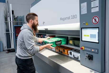 Mitarbeiter entnimmt einen grünen Werkzeugkoffer aus einem Kardex Megamat Essential. Die automatisierte Lagerlösung verfügt über einen integrierten Touchscreen zur Bedienung.